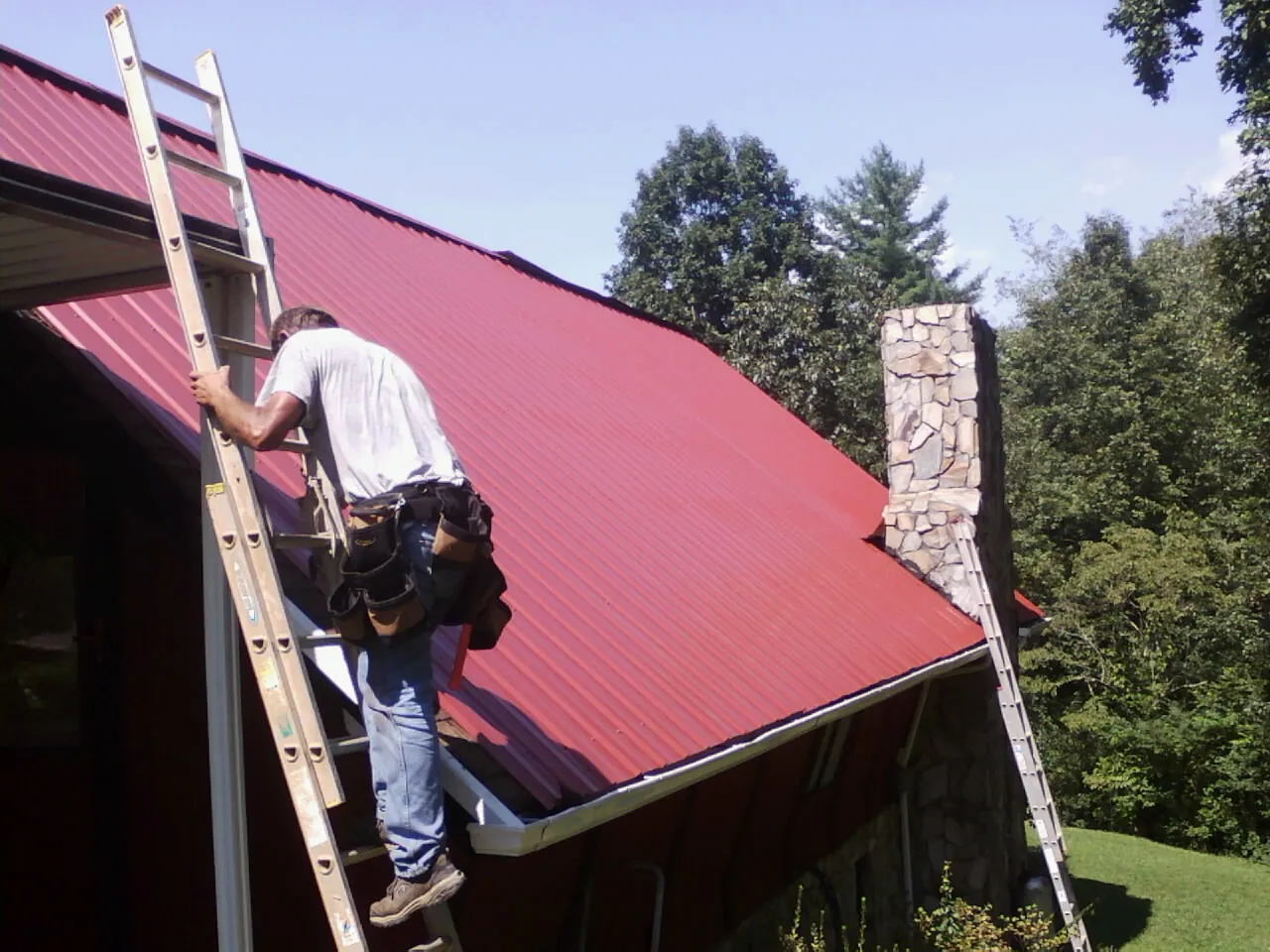 Roofer on a ladder beside a red metal roof and stone chimney — Soffit & Fascia Repair by Roofing Brothers in Allensville